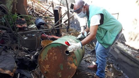 Homem usando colete de identificação, máscara de acetato e de tecido e luva, mexe em galão enferrujado. O recipiente, sem tampa, está em meio a diversas peças de ferro retorcidas. Há também algumas plantas. #Paratodosverem