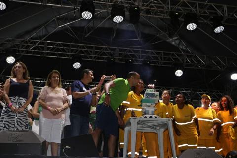 Hora de cantar o parabéns. Várias pessoas estão no palco próximas a um bolo cenográfico com o número 474 no topo. O prefeito e algumas margaridas que fazem a limpeza da praia sopram a velinha. #Pracegover