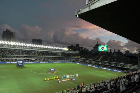 o gramado iluminado com os time em campo alinhados para cantar o hino nacional, no início do jogo. #paratodosverem