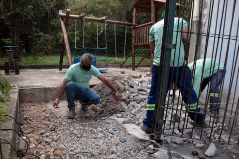 três homens estão em um pequeno cercado com o piso quebrado. Um deles segura uma trena estendida. #paratodosverem