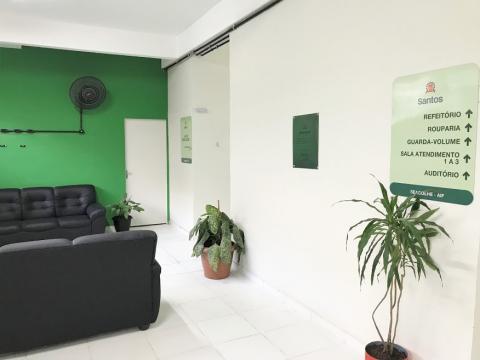 Sala de recepção branca e verde, com sofá, placas e vaso de plantas. #paratodosverem