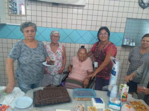 Senhoras e agente comunitária em pé ao lado de senhora em cadeira de rodas durante festa de aniversário. #pracegover