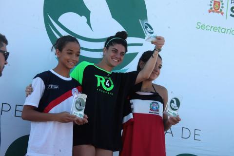Marcela Nicolau (centro da foto), Chiara Candido Paione (à esquerda) e Maria Eduarda de Oliveira Rodrigues (à direita), todas no pódio segurando troféus. #paratodosverem