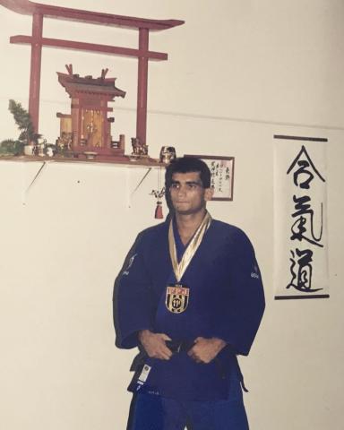 Imagem de Alex com quimono azul e medalha no pescoço com imagens da cultura japonesa ao fundo. #pracegover