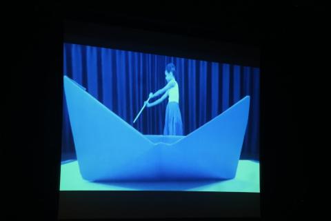 Vídeo no telão mostra menina navegando em um barco de papel #paratodosverem