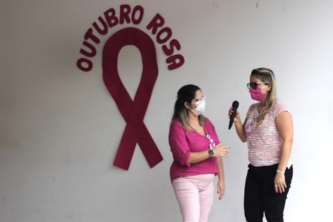 Duas mulheres, uma com o microfone e ao fundo a parede escrito outubro rosa #paratodosverem