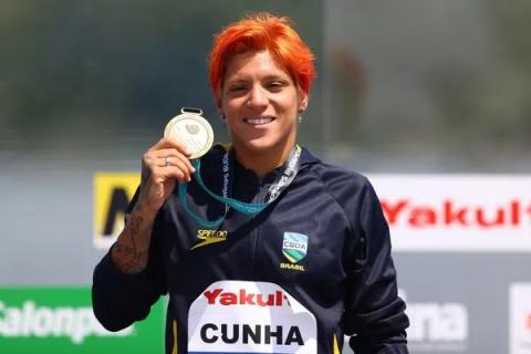 ana marcela segura a medalha com a mão direita ao lado do rosto. Ela sorri para a foto. #paratodosverem