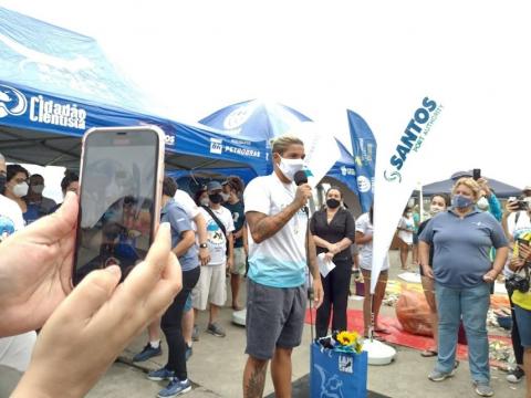 Ana Marcela Cunha fala ao microfone à frente de uma tenda. Em primeiro plano, mãos seguram um smartphone para fotografar a atleta. #paratodosverem