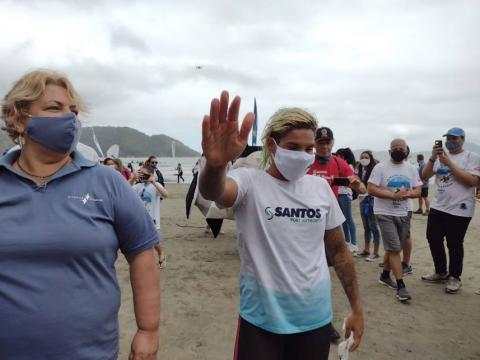 Ana Marcela caminha na areia, seguida por diversas pessoas, e acena para o público. #paratodosverem