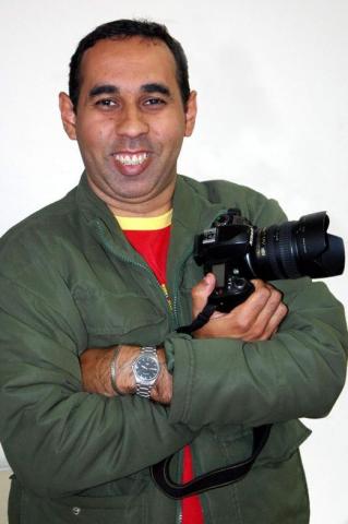 O fotógrafo, sorrindo com uma câmera na mão e braço cruzado. #pracegover