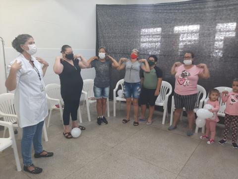 várias pessoas estão em pé fazendo movimentos com os braços. Uma mulher está usando avental, como se estivesse coordenando o grupo. #paratodosverem