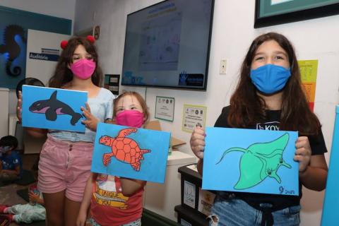 Três meninas exibem os quadros com pinturas de animais marinhos. #pracegover