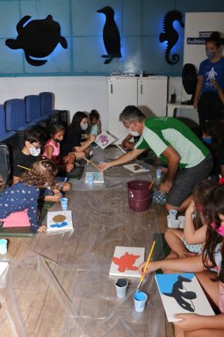 Crianças sentadas no auditório do parque pintam e são orientadas pelo arte-educador. #pracegover