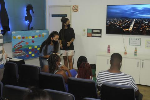 auditório com algumas pessoas sentadas olhando para uma tela onde está projetada uma imagem noturna de santos. Duas moças estão em pé, próximas a um painel alusivo a Santos. #paratodosverem