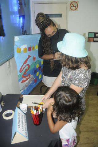 duas meninas estão junto a mesa se preparando para desenhar ou escrever. Elas falam com uma mulher que está diante do cartaz alusivo a Santos