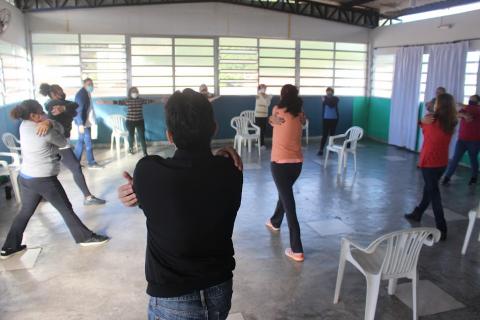 pessoas estão em pé se movimentando em um salão. Elas se autoabraçam num exercício de alongamento. #paratodosverem