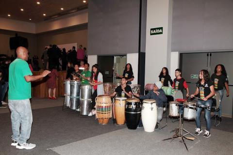 apresentação de músicos tocando instrumentos de percussão. Um homem à frente rege o grupo de jovens. #paratodosverem 