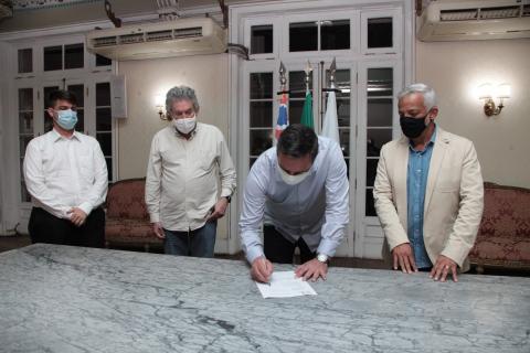 prefeito assina papel sobre uma mesa. Ele está ladeado por um homem à direita e dois à esquerda. #paratodosverem