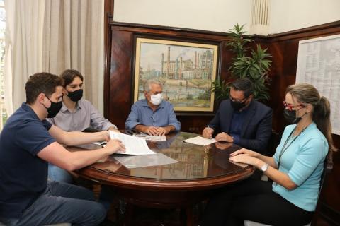 quatro pessoas sentadas em torno de uma mesa. O prefeito, o terceiro da esquerda para a direita, assina um papel. #paratodosverem