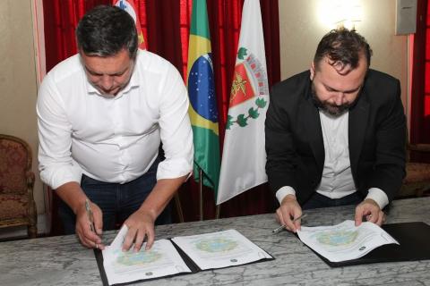 Prefeito e superintendente da SPU assinam documento sobre uma mesa. Bandeiras de Santos e do Brasil estão visíveis atrás. #paratodosverem