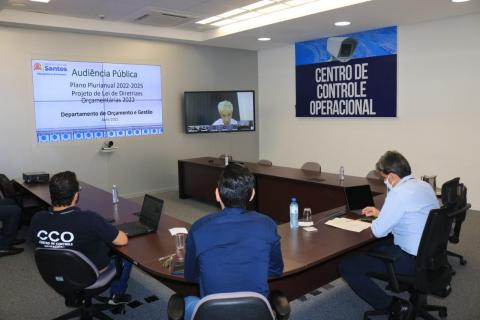 reunião com três pessoas em caráter presencial em torno de uma mesa e uma outra, no telão ao fundo. #paratodosverem