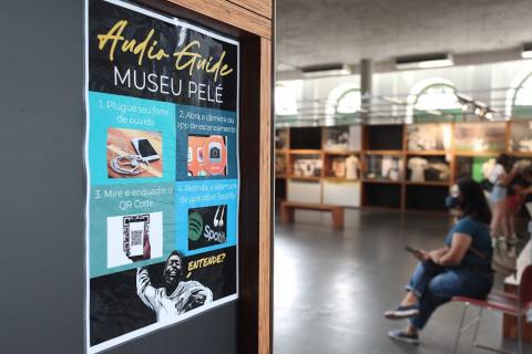 porta de entrada com cartaz afixado onde se lê Audio Guide Museu Pelé. O carta tem algumas orientações para o funcionamento do serviço, com uma imagem de pelé comemorando um gol na parte de baixo. #paratodosverem