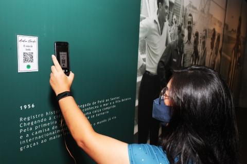 mulher está em pé apontando celular para QR code afixado em parede ao lado de foto em tamanho gigante. #paratodosverem