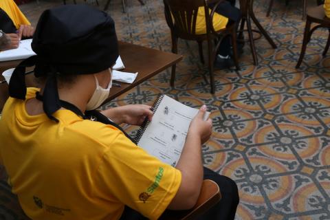 Aluno usando uniforme e sentado, lê capa de apostila do curso. #paratodosverem