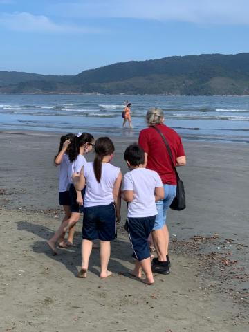 alunos caminham descalços na areia, acompanhados de uma adulta. #paratodosverem