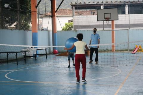 quadra esportiva com cesta de basquete ao fundo. No centro uma rede e uma menina de costas segurando uma bola grande. Do outro lado, um homem e uma criança. #paratodosverem