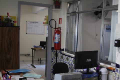 extintor está preso a parede próximo à janela de uma sala. Há um computador e papéis sobre a mesa em primeiro plano. #paratodosverem