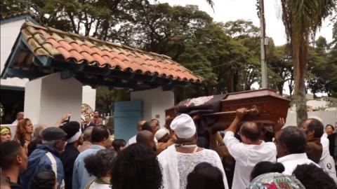 Pessoas acompanham trajeto de caixão que está sendo carregado. Cena de Axexe - #paratodosverem