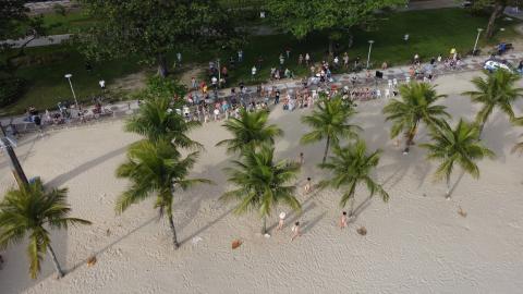 Foto aérea mostra bailarinas na areia, coqueiros na praia e pessoas assistindo #paratodosverem