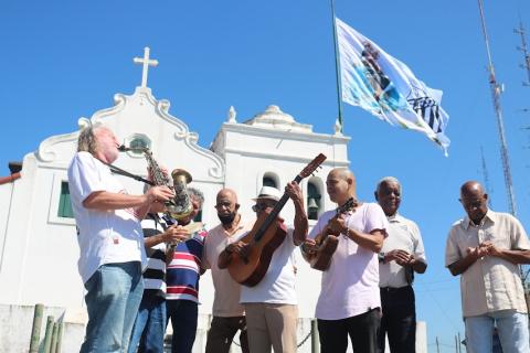 músicos com instrumentos tocam em frente à capela e abaixo da bandeira hasteada. #paratodosverem