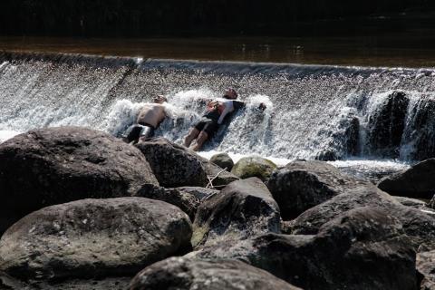 pequena cachoeira com dois homens recostados e de braços abertos sendo banhados. Pedras em primeiro plano. #paratodosverem