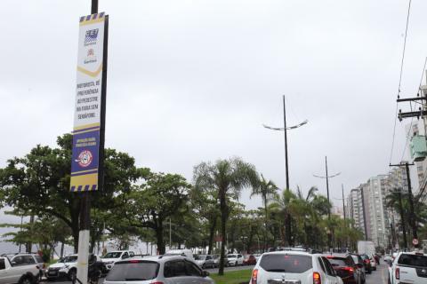 banner afixado em poste em canteiro central de avenida da orla. Carros passam nas duas pistas, indo e vindo. #paratodosverem