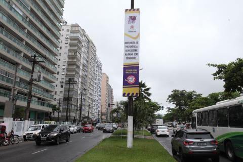 banner afixado em poste em canteiro central de avenida da orla. Carros passam nas duas pistas, indo e vindo. #paratodosverem