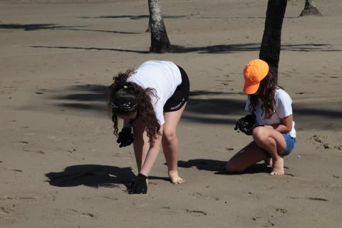 duas meninas estão agachadas na areia recolhendo microlixo. #paratodosverem