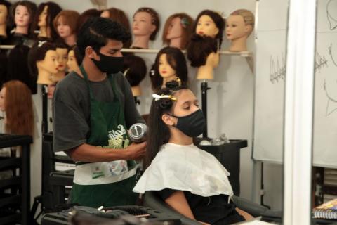 homem faz escova no cabelo de uma mulher. Ao fundo, várias manequins de cabeça com perucas. #paratodosverem