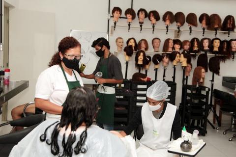 mulher está sentada, de costas para a foto, com o cabelo preparado para o tratamento. Uma mulher à frente dele está fazendo o serviço de manicure. #paratodosverem