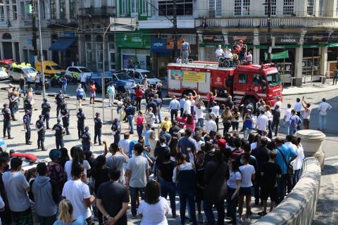 Praça com várias pessoas e ao fundo carro do corpo de bombeiros #paratodosverem