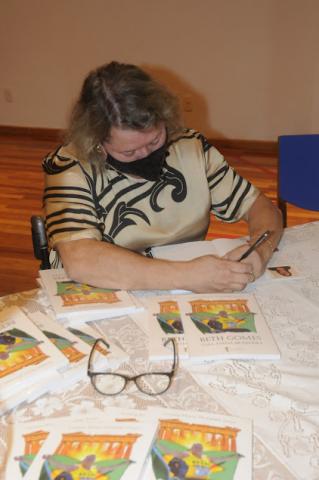 Beth está sentada atrás de uma mesa autografando o livro. Hà várias publicações sobre a mesa. #paratodosverem