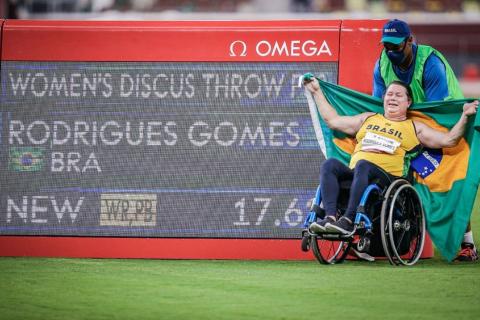 Na cadeira de rodas, Beth é conduzida para dentro de gramado segurando a bandeira do Brasil. Ao lado, um painel com o nome dela. #paratodosverem