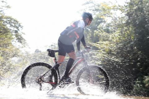 ciclista percorre trecho de rio. Água espirra por vários lados. #paratodosverem