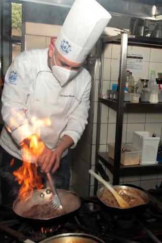 Chef de cozinha prepara refeição com labareda saindo de frigideira durante fritura. #pracegover