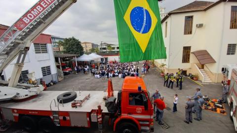carro de bombeiro com escada magirus elevada e bandeira do brasil pendurada. Ao fundo as crianças em um pátio. #paratodosverem
