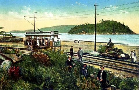 bonde na orla de santos, com vegetação em primeiro plano, a linha do bonde, e uma pequena ilha, no meio do mar, ao fundo, provavelmente a Ilha de Urubuqueçaba. #paratodosverem