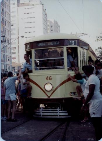 bonde circula pela orla, lotado de passageiros e pessoas do lado de fora. #paratodosverem