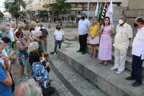 Pessoas estão reunidas na praça. Representantes da cidade e instituições estão num patamar mais alto falando ao público, abaixo. #paratodosverem