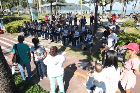 crianças e adolescentes estão sentados em cadeiros fazendo apresentação de flauta. Eles estão de costas para o jardim. O público assiste. #paratodosverem